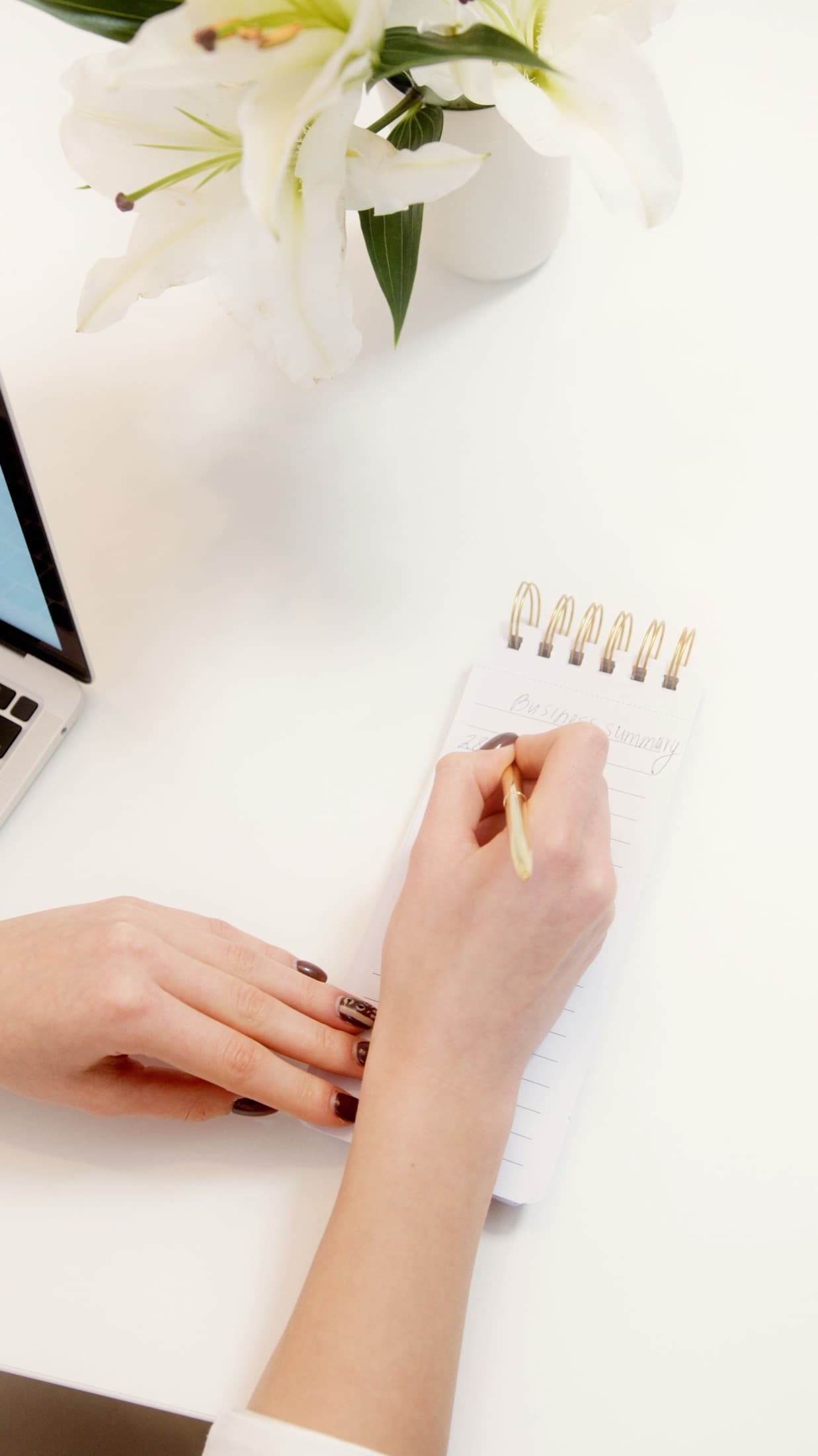 Main qui écrit sur un carnet à spirale, bureau clair et lys blancs